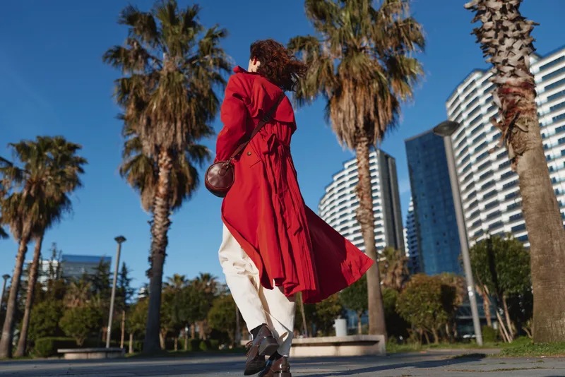A young woman in a flowing red trench coat walks confidently along a sunny urban path lined with palm trees and modern buildings.