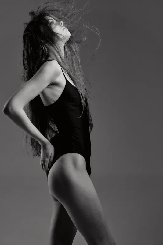 Black and white artistic portrait of a slender woman in a bodysuit, expressing confidence and grace with dynamic hair movement.