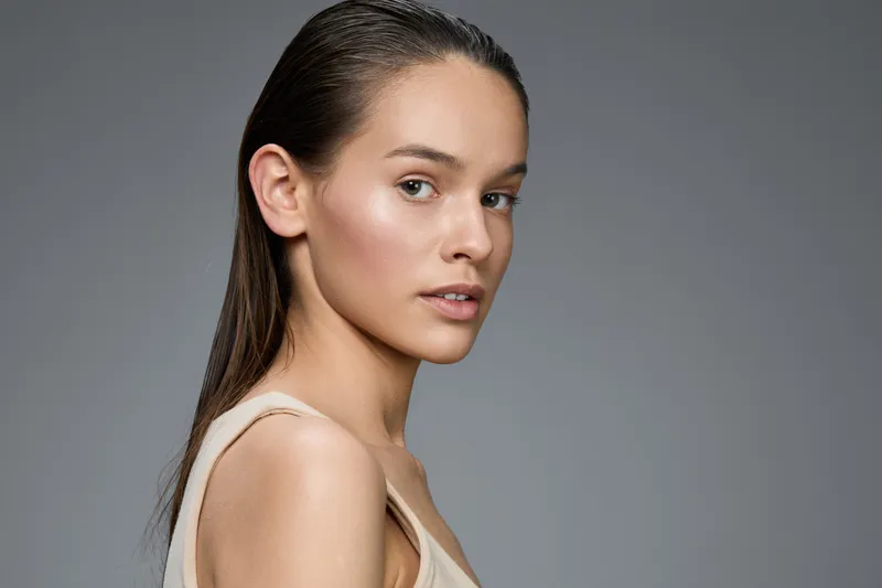 Portrait of a young woman with long hair looking confidently at the camera, featuring natural makeup and a neutral background, ideal for beauty and skincare concepts