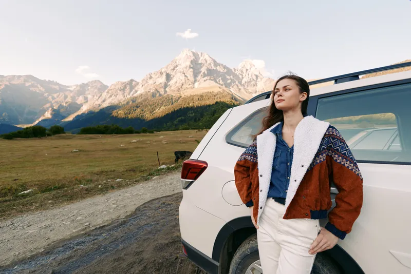 Woman SUV mountains landscape travel adventure portrait outdoor scene. Casual young adult leaning on car in denim jacket, confident roadtrip explorer enjoying serene scenic horizon and freedom.
