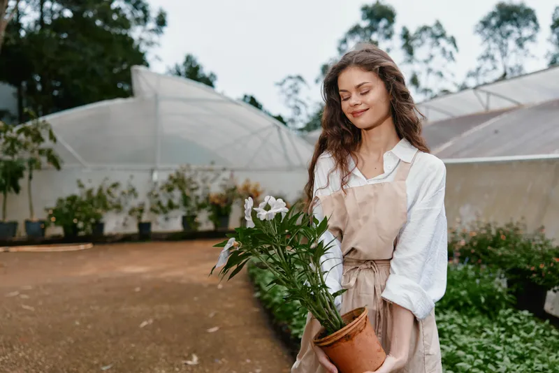 Young woman in a garden holding a potted flower, wearing an apron, smiling, showcasing a love for gardening and nature in a serene setting