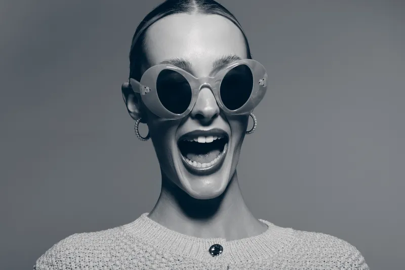 Happy caucasian woman with light skin wearing round sunglasses and hoop earrings smiling widely in studio. Concept for fashion, joy, and positive emotions.