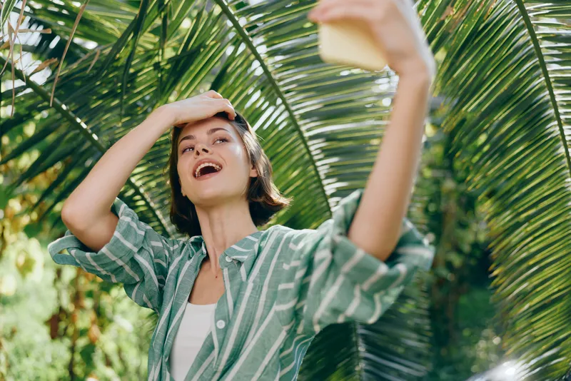 Joyful outdoor portrait of a woman shielding eyes from the sun while reaching for a selfie among lush palm leaves and bright tropical light.