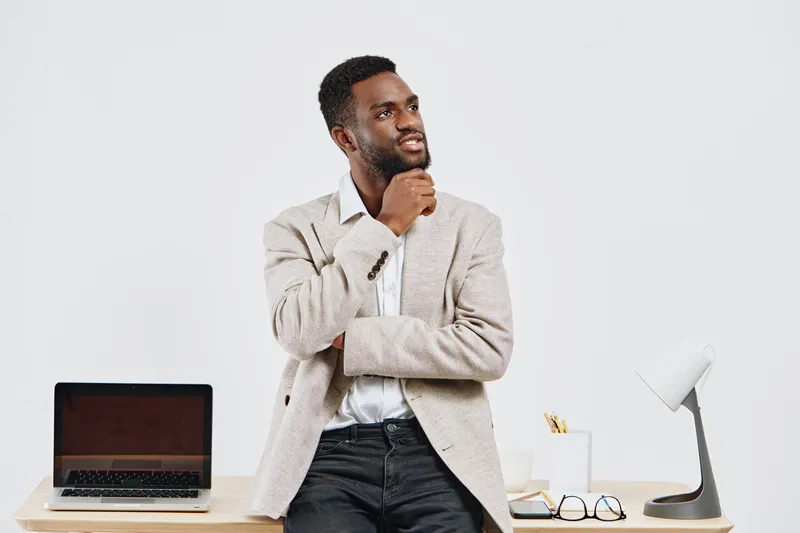 Thoughtful smiling man in beige blazer and white shirt standing at desk with laptop and glasses in modern office interior, contemplating idea or decision, business casual style, professional atmospher