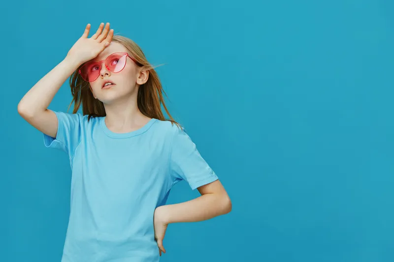 Young girl in a light blue t shirt and pink sunglasses, posing with a playful expression against a bright blue background, capturing summer vibes and fun emotions