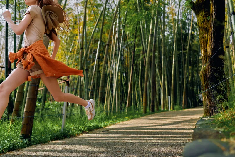 Girl jumping on a forest path with a backpack among tall bamboo, summer motion captured midair, carefree outdoor travel and energetic leap on a sunny nature trail.
