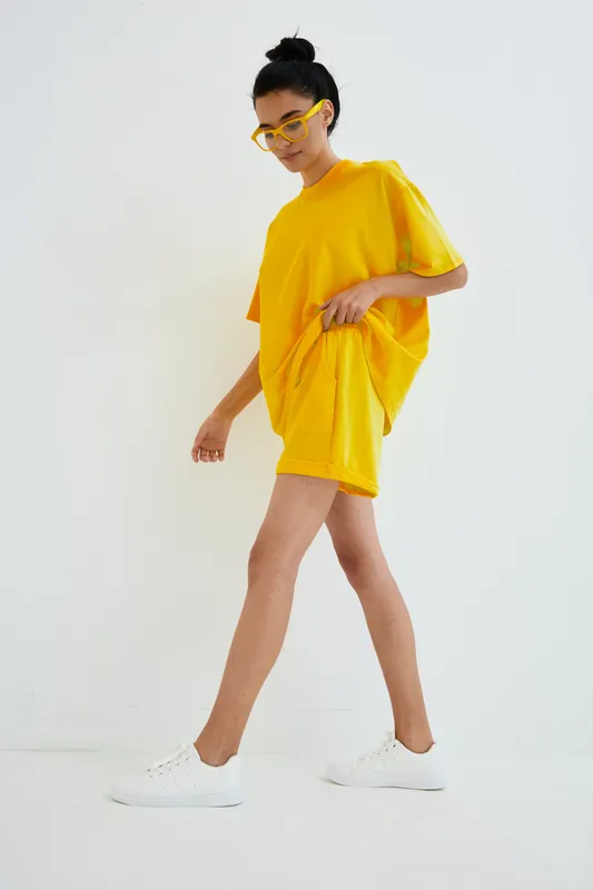 Young woman walking confidently in bright yellow casual outfit with matching glasses and white sneakers on light minimalist background.