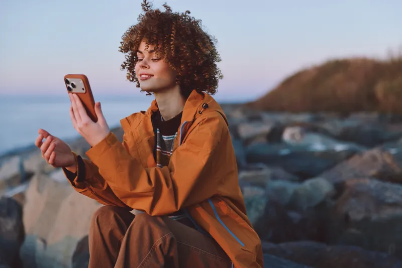 Woman lifestyle outdoors, curly hair orange jacket, smiling with smartphone on rocky beach, sea waves and sunset sky, casual pose, natural light