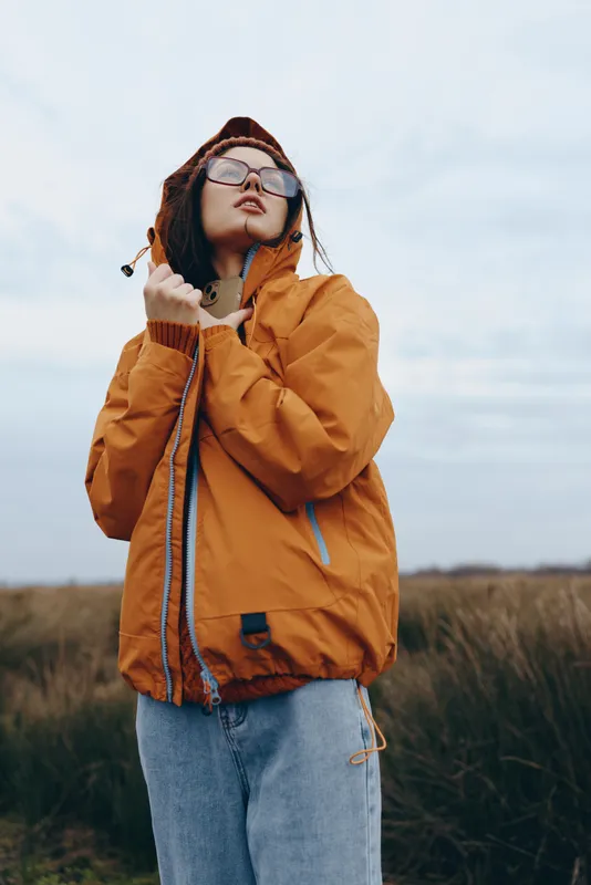 Woman lifestyle outdoors in field, orange jacket, sunglasses on face, headphones, listening while looking upward, calm relaxed pose, cloudy sky, grass horizon, fresh air, gentle breeze