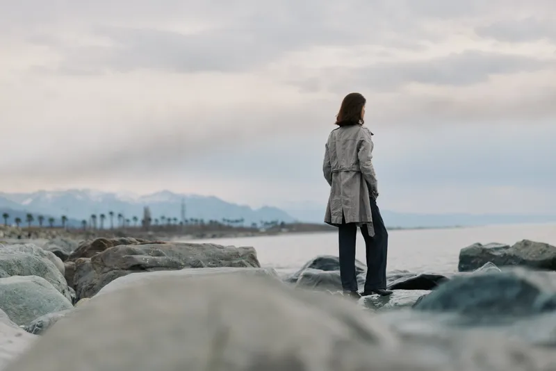 A woman in a coat stands on rocks by the sea, looking at distant mountains under a cloudy sky. The scene evokes solitude, nature, and peaceful reflection at the coastline.