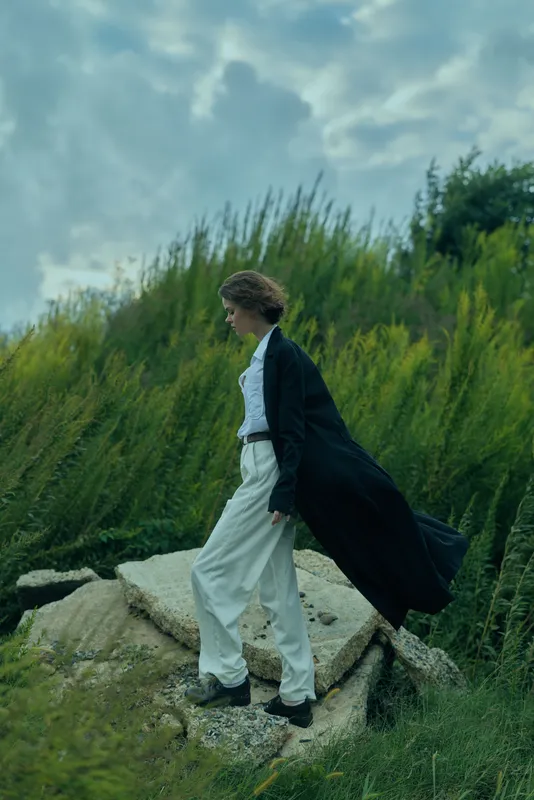 Woman in a long black coat walks along rugged rocks, white suit visible beneath, wind sweeping grass and moody sky creating a dramatic outdoor fashion moment
