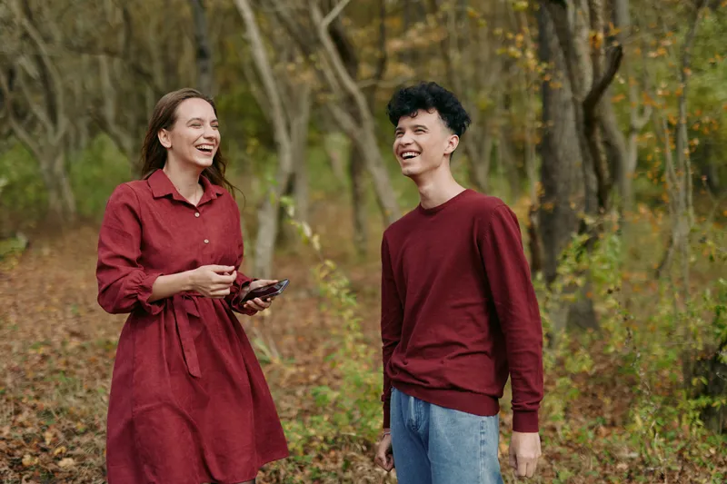 Two friends share a candid moment outdoors in an autumn forest, displaying genuine smiles and authentic interaction that convey warmth and believable connection.
