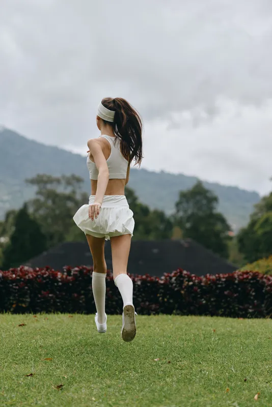 Athletic young woman in white sportswear running joyfully across green grass with mountains in the background, showcasing a healthy lifestyle and fitness concept
