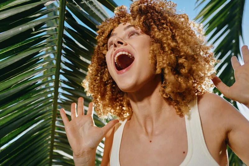 smiling woman with curly hair enjoying tropical sunlight behind palm leaves, expressing joy and freedom in summer outdoors vibrant mood