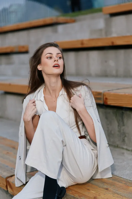 Young white woman in stylish white outfit sitting on wooden stairs looking away confidently with neutral gray urban background, autumn fashion concept