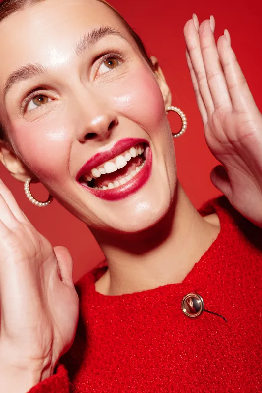 Happy woman with bright red lipstick and hoop earrings smiles widely, wearing a red sweater against a red background. Concept of joy, fashion, beauty, and vibrant style.