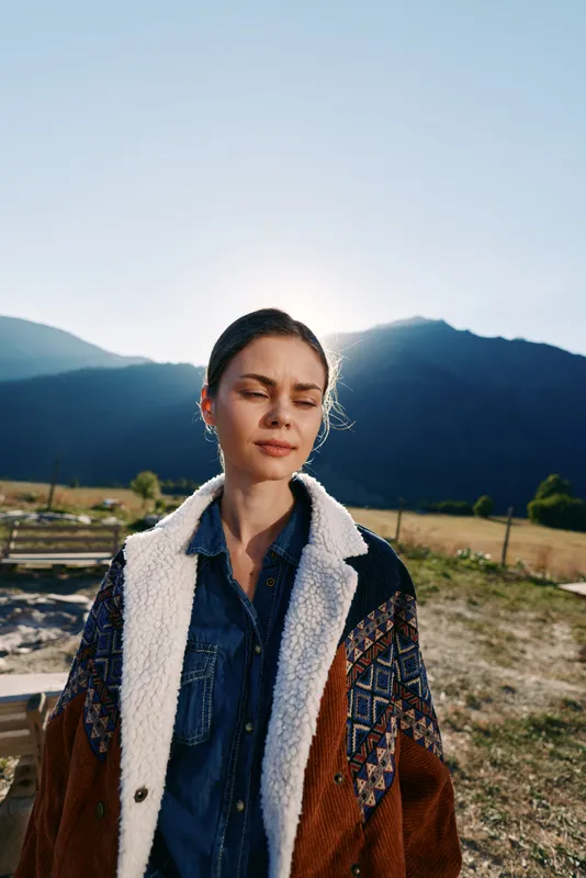 Woman portrait in mountains with sunlight behind her, eyes closed and serene expression, wearing warm patterned coat and denim shirt in outdoor nature with soft backlight.