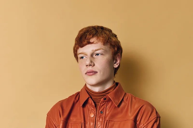 Young man with red hair and fair skin wearing brown jacket standing against plain light brown background looking sideways with calm expression and relaxed posture.