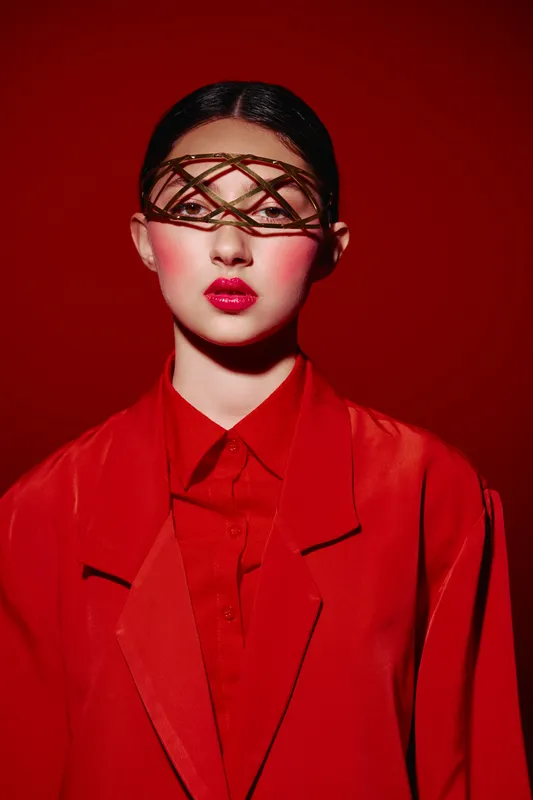 Fashion model in vibrant red coat and shirt faces camera with elaborate decorative brow accessory framing eyes. Studio portrait with intense red background. Concept of luxury beauty and fashion.