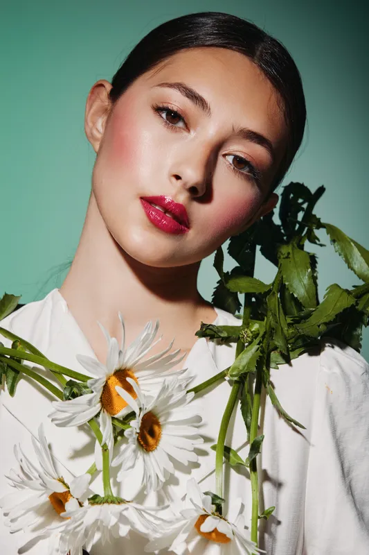 Young woman with glossy red lips and radiant makeup wears white flowers on her shoulder. Soft green background. Creative beauty and fashion portrait with vibrant colors.