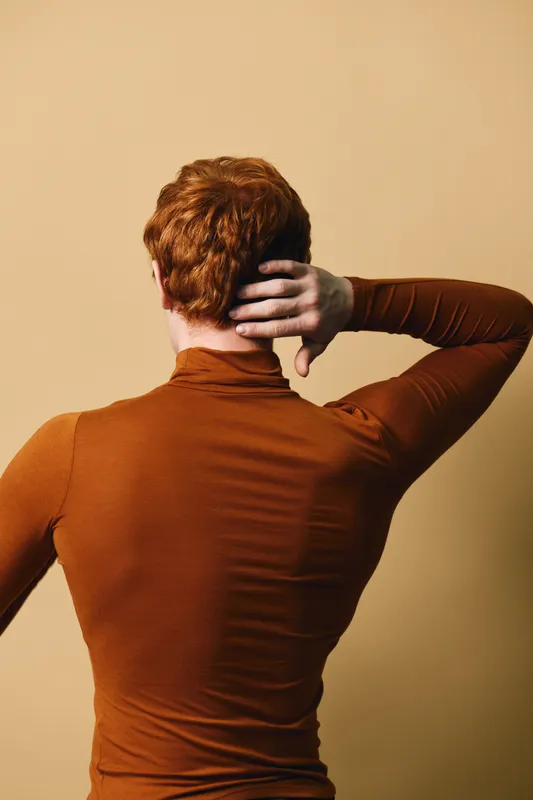 Young man with red hair wearing brown long sleeve stands against plain beige wall touching back of head with hand. Casual posture showing back view with minimalistic warm colors.