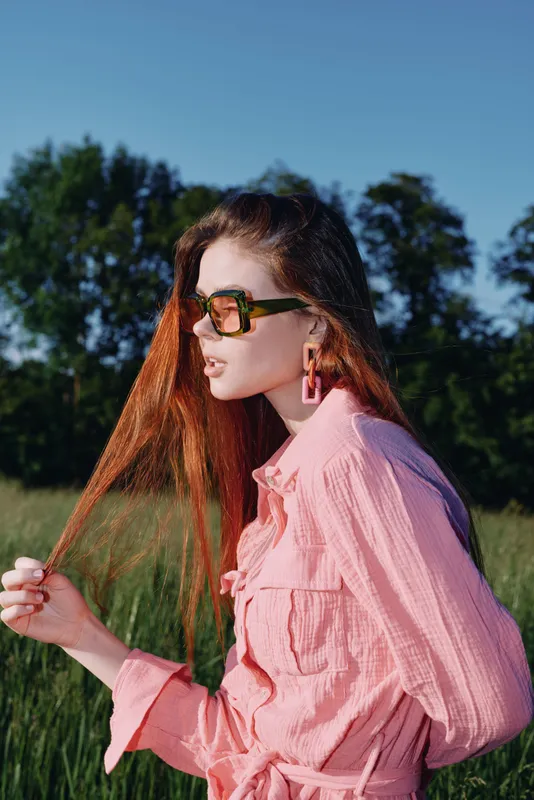 Stylish woman with long red hair wearing a pink shirt and sunglasses in a sunny field, showcasing confidence and summer fashion trends