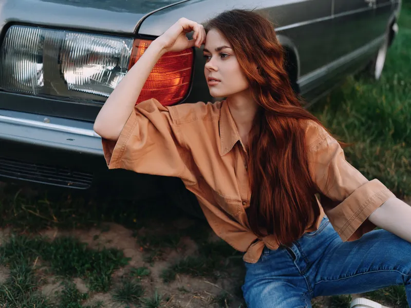 Young woman with long red hair sitting by vintage car in casual brown shirt, expressing a thoughtful mood in a green outdoor setting.