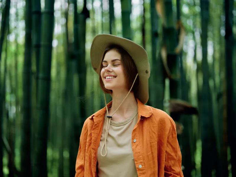 Woman smile hat bamboo forest outdoors nature portrait of a young female in orange jacket, eyes closed and peaceful, joyful traveler relaxing amid greenery and soft sunlight in nature.