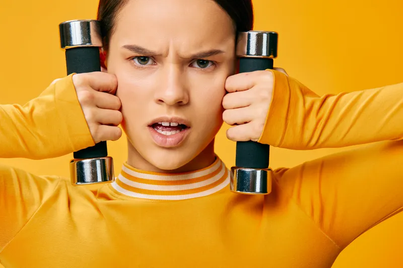 Focused woman in bright orange athletic wear grips dumbbells near her face, displaying strength and determination during a high-energy fitness workout against a vivid yellow backdrop.