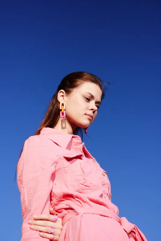 Stylish young model in a pink outfit with colorful earrings against a clear blue sky, embodying confidence and modern fashion trends