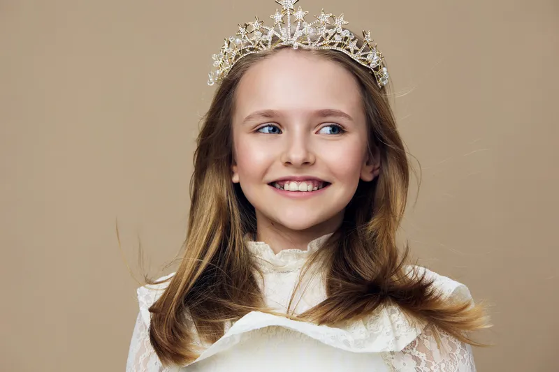 Young girl with a crown smiles joyfully, wearing a beautiful lace dress, showcasing elegance and innocence with a neutral background, perfect for children s photography