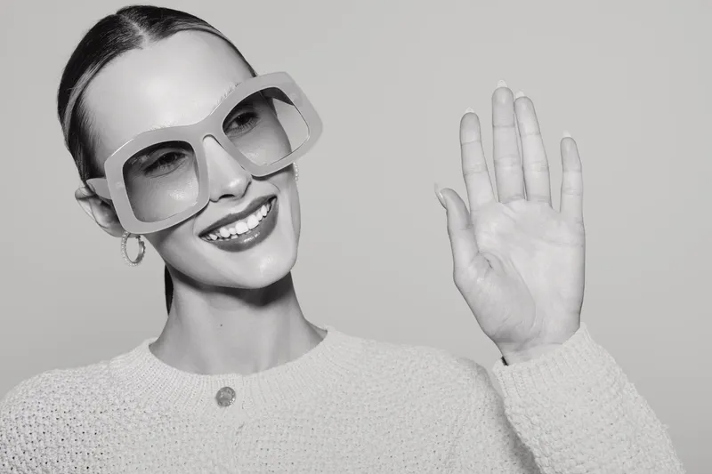 Smiling caucasian woman with fair skin wearing large square sunglasses waves hand. Modern fashion style, positive emotion, casual outfit, isolated on plain background concept.