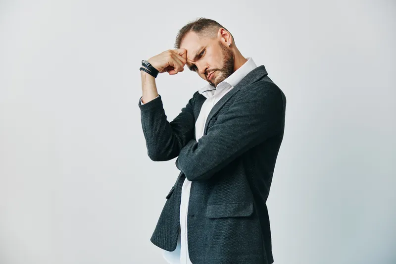 A focused business professional stands against an isolated colored background, conveying thoughtful introspection and resolve as he rests his head on a hand.
