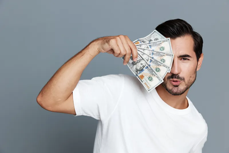 Confident man holding cash bills close to face, looking at camera with playful expression. Minimalistic background highlights the joyful moment of celebrating success.