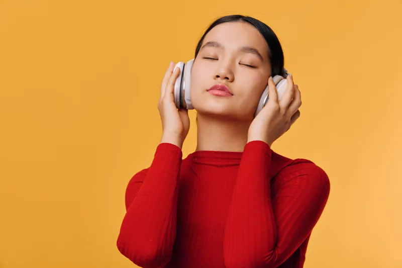 Young woman wearing headphones and closed eyes enjoying music with calm face, standing against yellow background in casual red sweater for relaxation and entertainment.