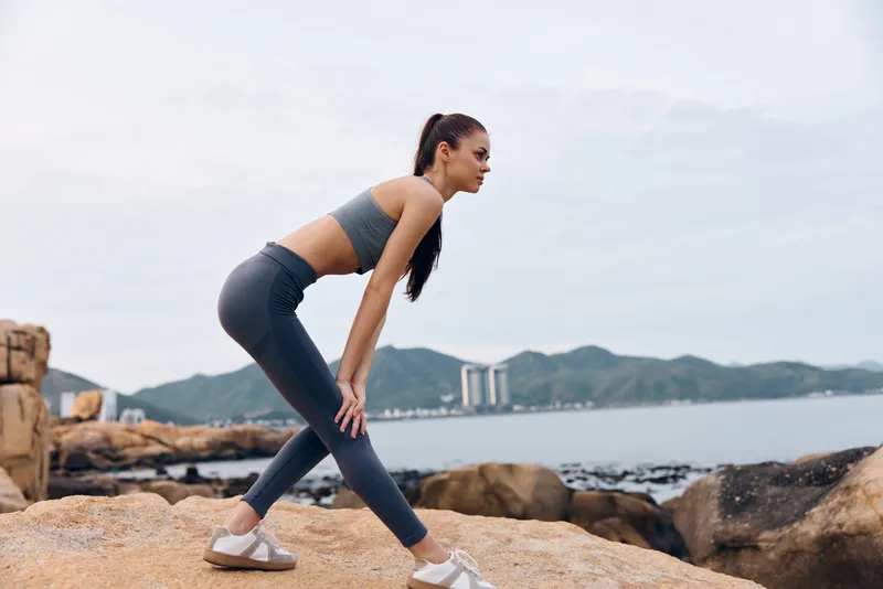 Young athletic woman stretching in sportswear on a rocky beach with an ocean view, promoting a healthy lifestyle and fitness attitude