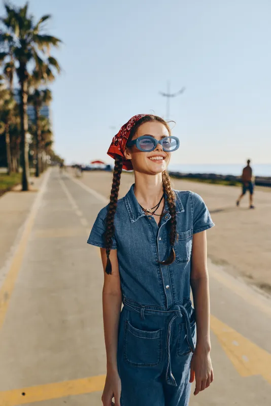 Lifestyle woman in modern boho-western denim street style with braided hair and blue sunglasses, smiling outdoors on a sunny day, film color effect enhancing natural tones.
