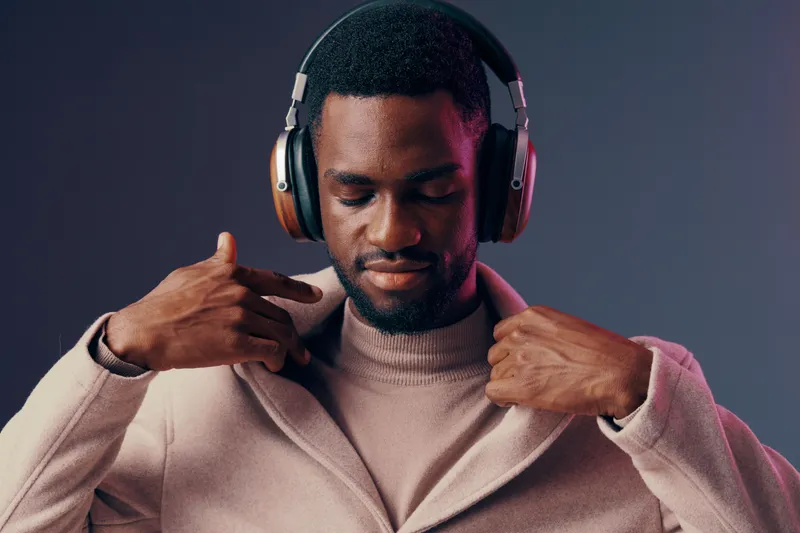 Stylish young Black man in headphones enjoying music, wearing a trendy coat against a gradient background, expressing calmness and confidence