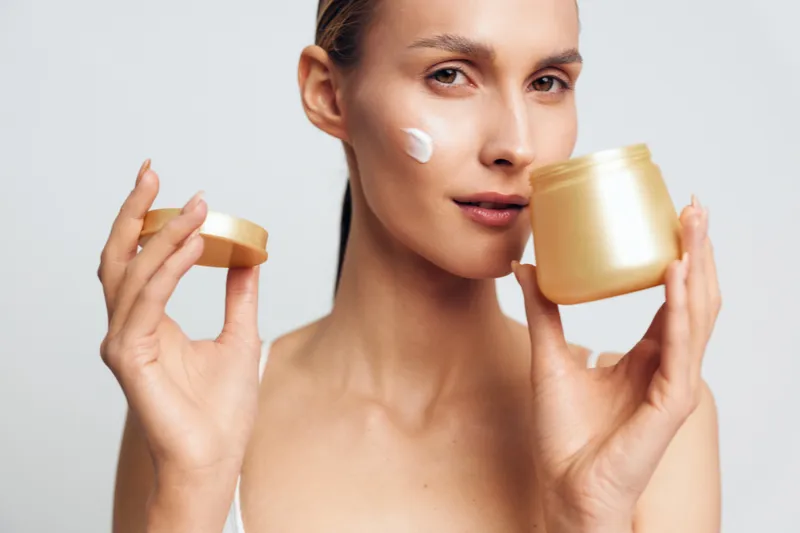 Creamy moisturizer applied on the cheek by a woman, holding the jar, showcasing a simple skincare routine, smooth skin and a healthy glow, studio portrait, closeup texture