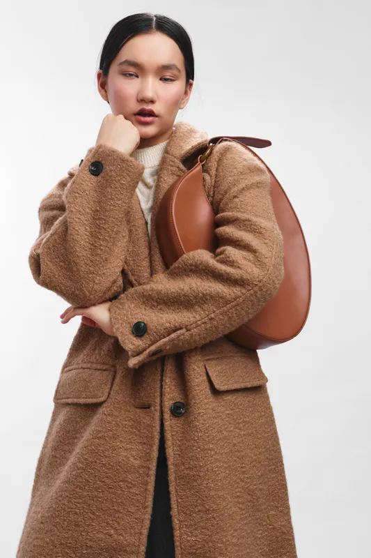 asian woman wearing a warm brown coat with a leather backpack poses in a studio, exuding confidence and style in a minimal fashion portrait.
