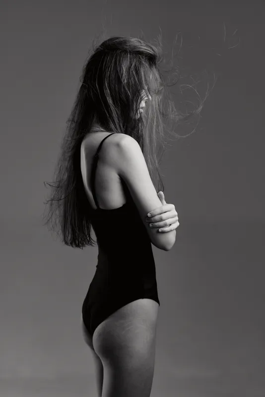 Black and white portrait of a slender woman with long hair in a bodysuit standing sideways with arms crossed, expressing calm and introspection.