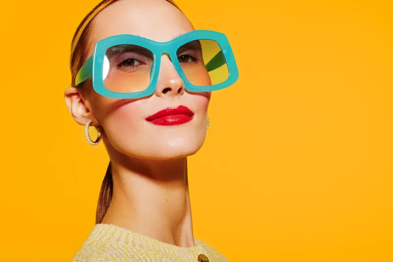 Confident woman with bright red lipstick and large turquoise glasses poses against vibrant yellow background. Fashion portrait with bold colors and modern style concept.