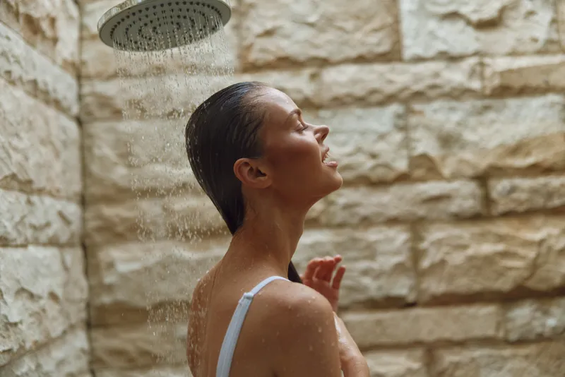 woman showering outdoors with closed eyes under rain showerhead, wet hair, stone wall background, natural light, serene mood, summer wellness moment