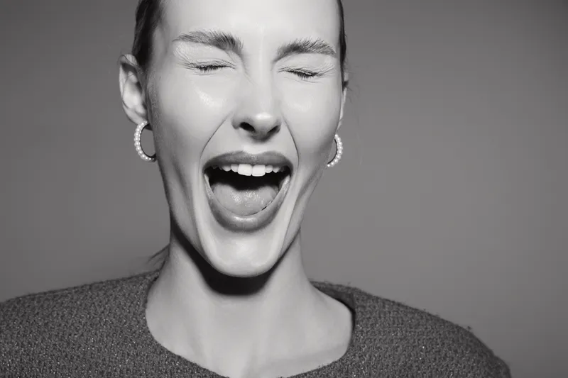 Young white woman with pale skin wearing earrings and knitted sweater laughing with closed eyes in black and white portrait.