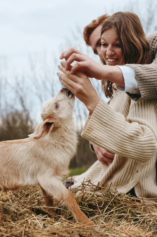 A young woman in a beige sweater feeds a baby goat outdoors while smiling and enjoying a natural lifestyle moment with another person in the background.