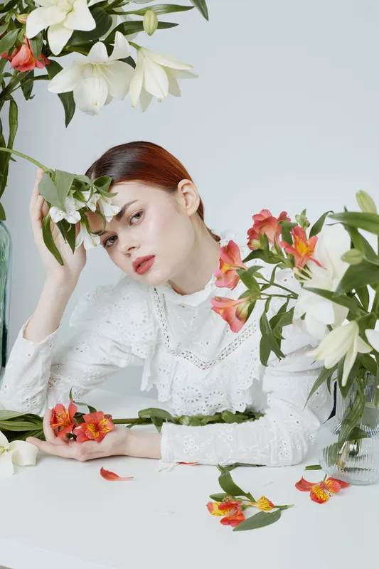 Young woman with red hair in white lace dress surrounded by colorful flowers, expressing contemplation and calmness, perfect for seasonal beauty themes