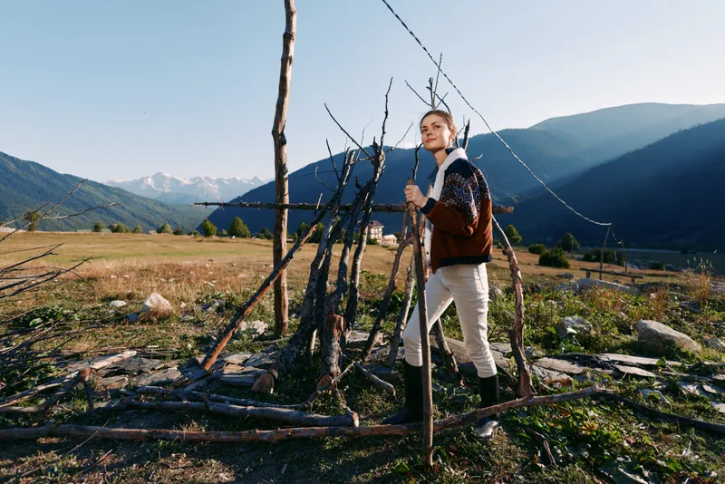 Woman hiking in mountains and nature across a rural meadow, walking with a wooden stick near a rustic fence. Portrait travel scene with sunlight, valley landscape and peaceful exploration.