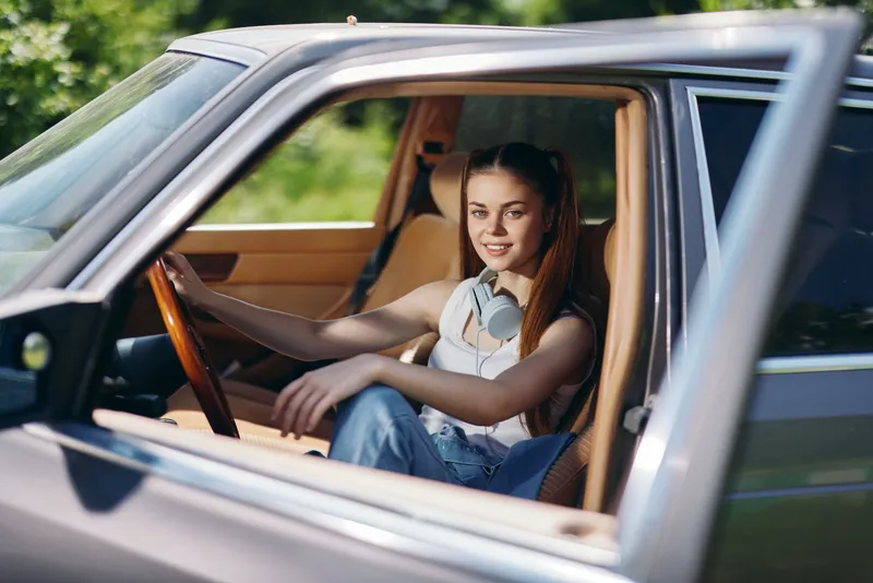 Young woman with headphones in a vintage car, smiling, dressed casually in a sleeveless top and jeans, surrounded by greenery in a bright, sunny setting.