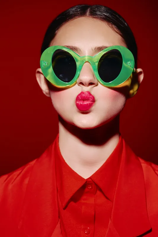 Young woman in oversized green sunglasses and bright red lips makes a kiss face. Bold makeup and red shirt against red background create a striking beauty fashion concept.