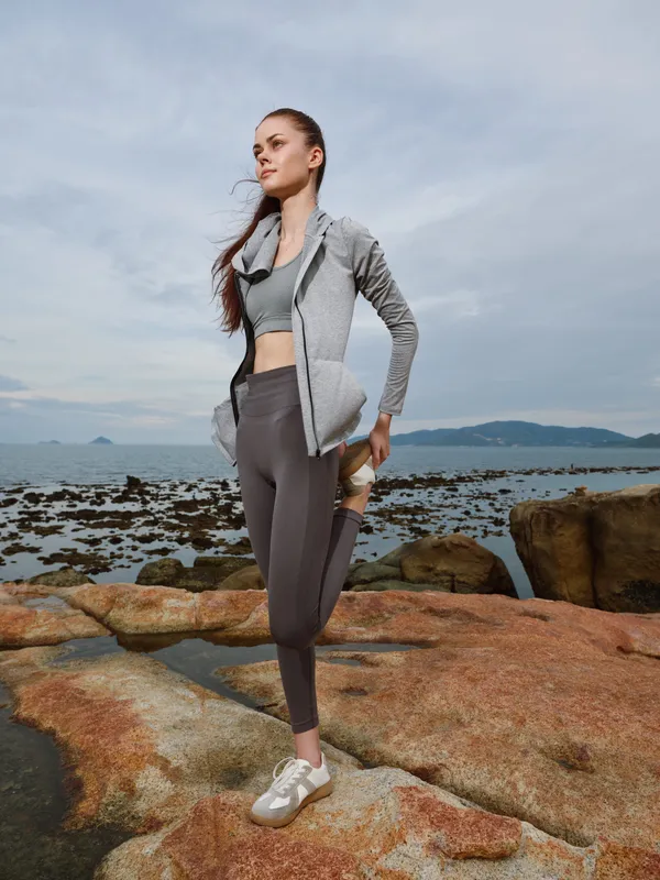 Stylish athletic woman in sporty outfit stretching by the sea at sunset, embodying strength and wellness, perfect for health and fitness themes